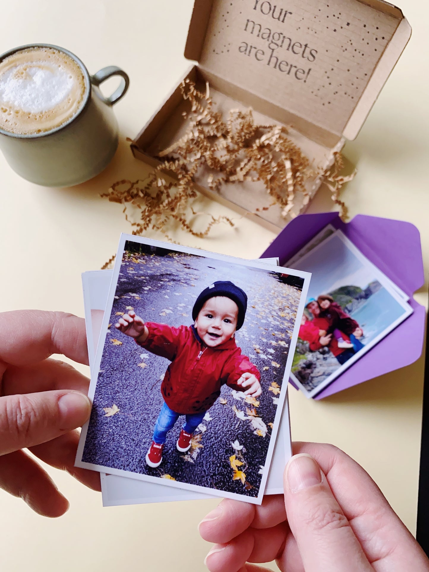 Hand holding a custom photo magnet above an open Magnetic Love gift box, with a coffee cup on a beige table.