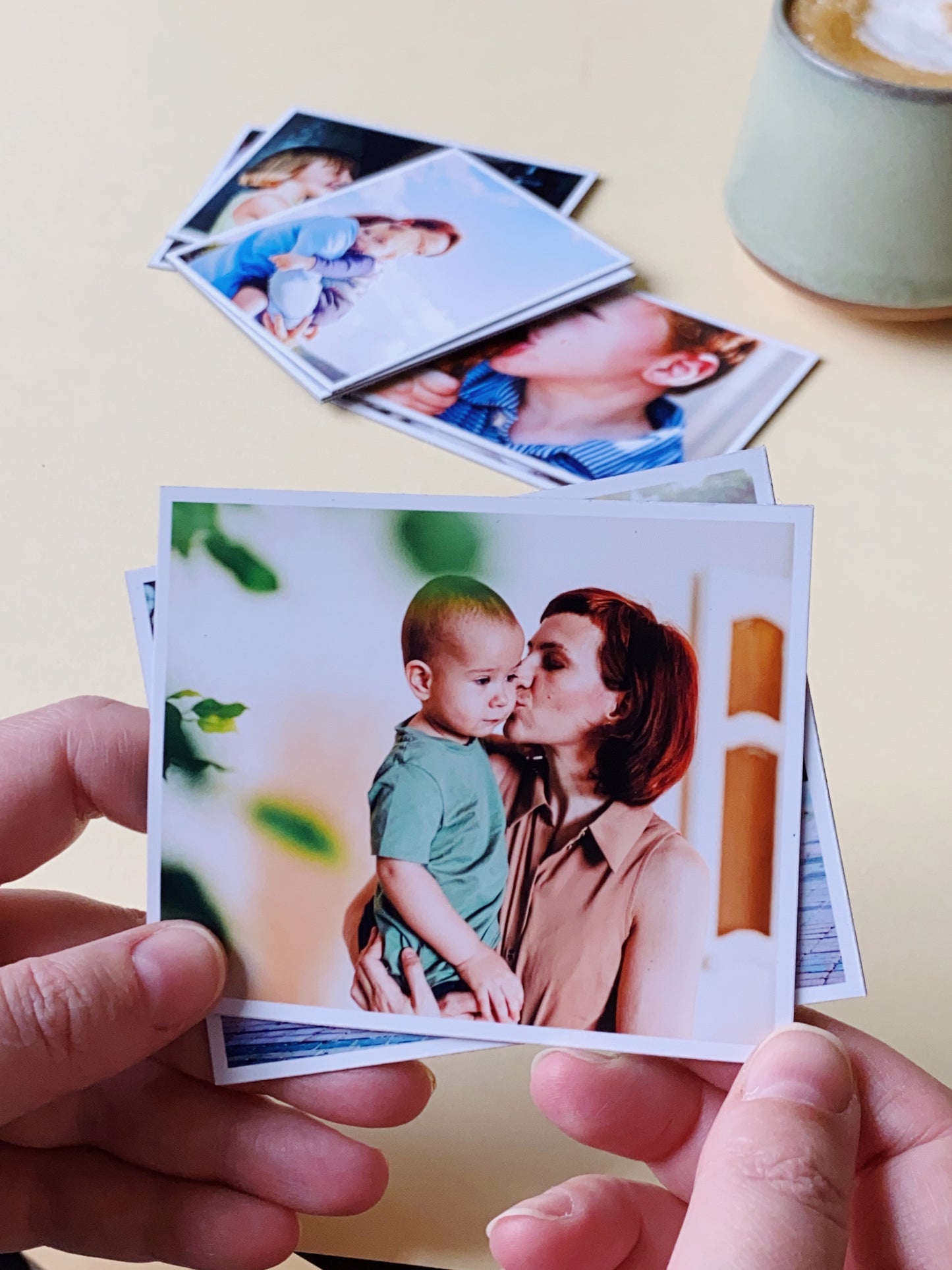 Hand holding a square custom photo magnet showing a mother kissing her child, with printed photos in the background.
