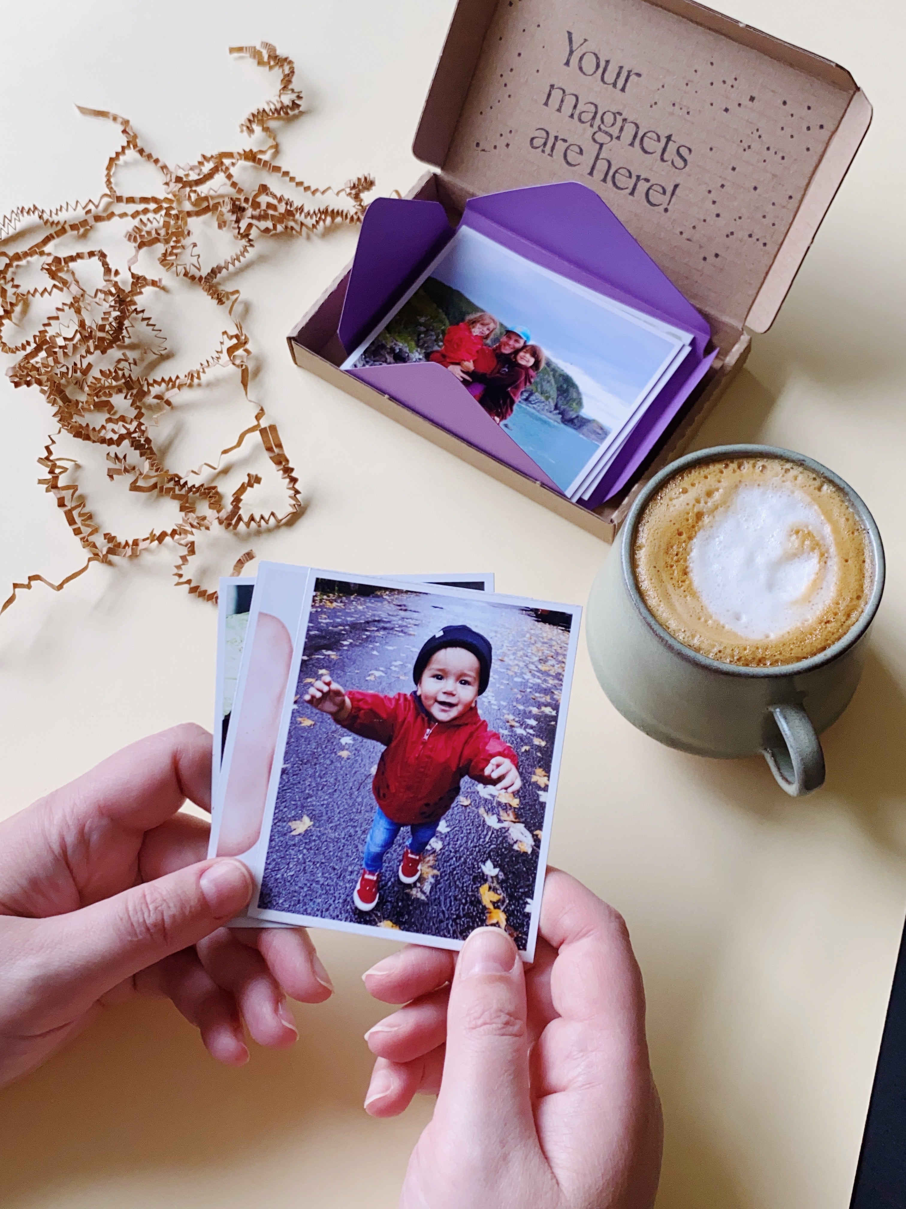 Hand holding a custom photo magnet above an open Magnetic Love gift box with a coffee cup on the table.